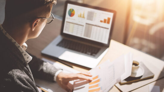 Young finance market analyst in eyeglasses working at sunny office on laptop while sitting at wooden table.Businessman analyze document in his hands.Graphs and diagramm on notebook screen.Blurred.