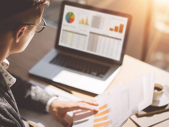 Young finance market analyst in eyeglasses working at sunny office on laptop while sitting at wooden table.Businessman analyze document in his hands.Graphs and diagramm on notebook screen.Blurred.